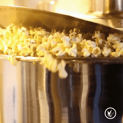 Fluffy yellow popped corn kernels push up the lid and overflow from a huge silver pot