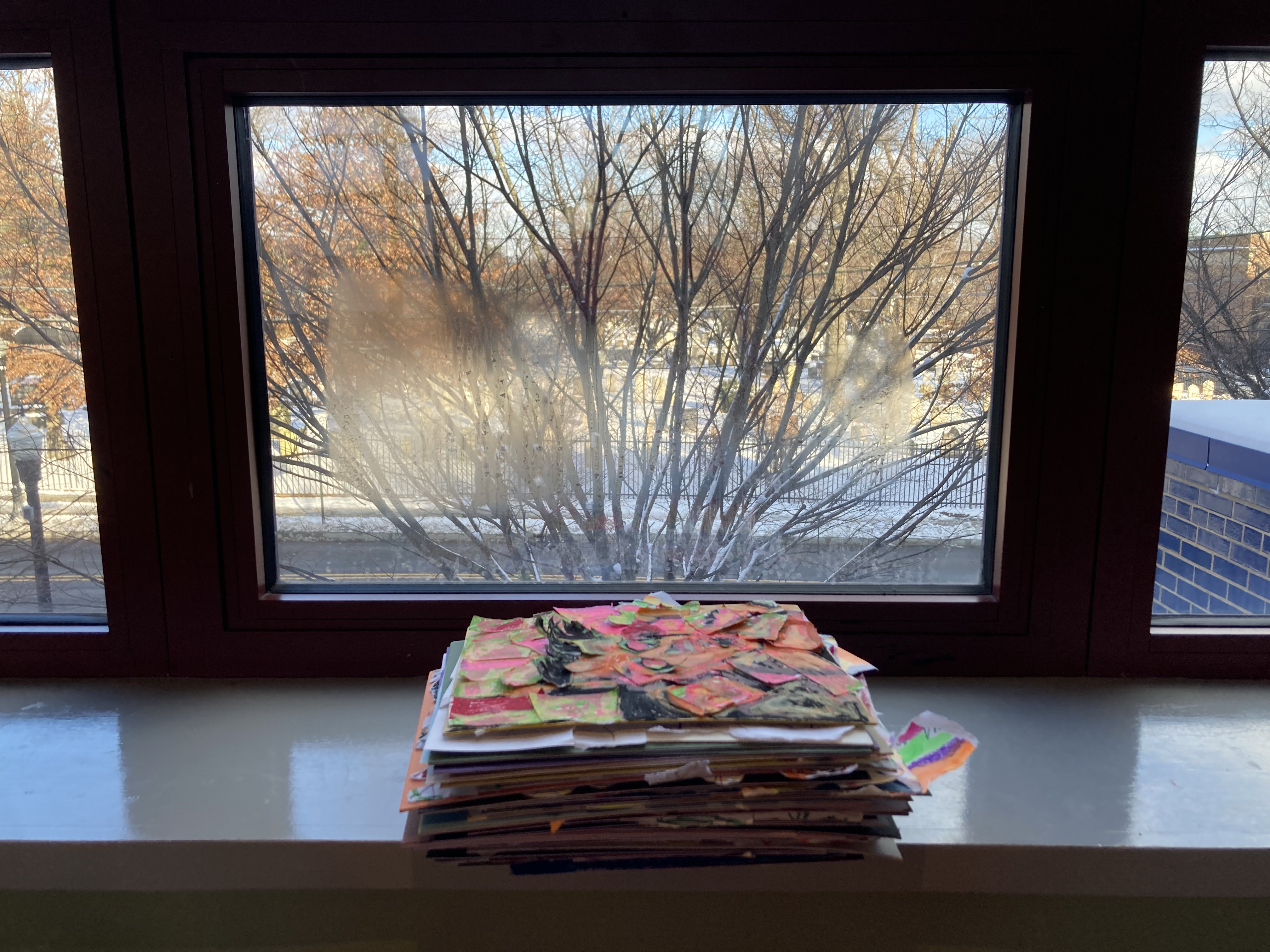 Dozens of collages stacked neatly on a shiny windowsill. The jagged bright scraps of paper that stick out from the pile are a nice contrast to the frosted window, bare branches, and snow outside.