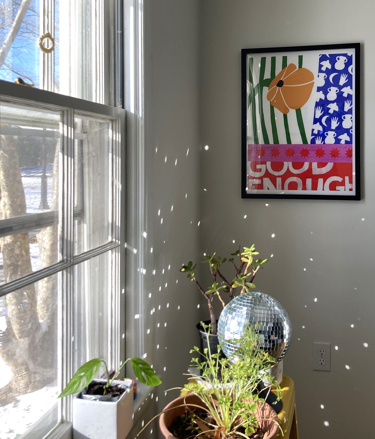 Sun streaming through my dining room window. There's a blue sky outside and snow on the ground. AN Anisa Makhoul poster is framed on the wall and reads GOOD ENOUGH in words that are legible but intentionally cut off. Some plants sit on a stool in the foreground, along with a disco ball that's casting prismatic light around the corner of the room.