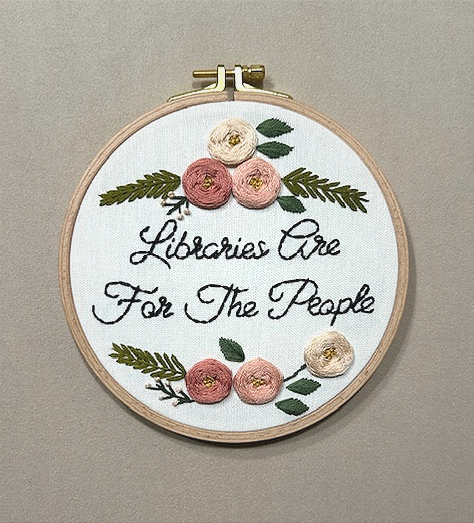 A needlepoint hoop with a floral design and the words LIBRARIES ARE FOR THE PEOPLE.
