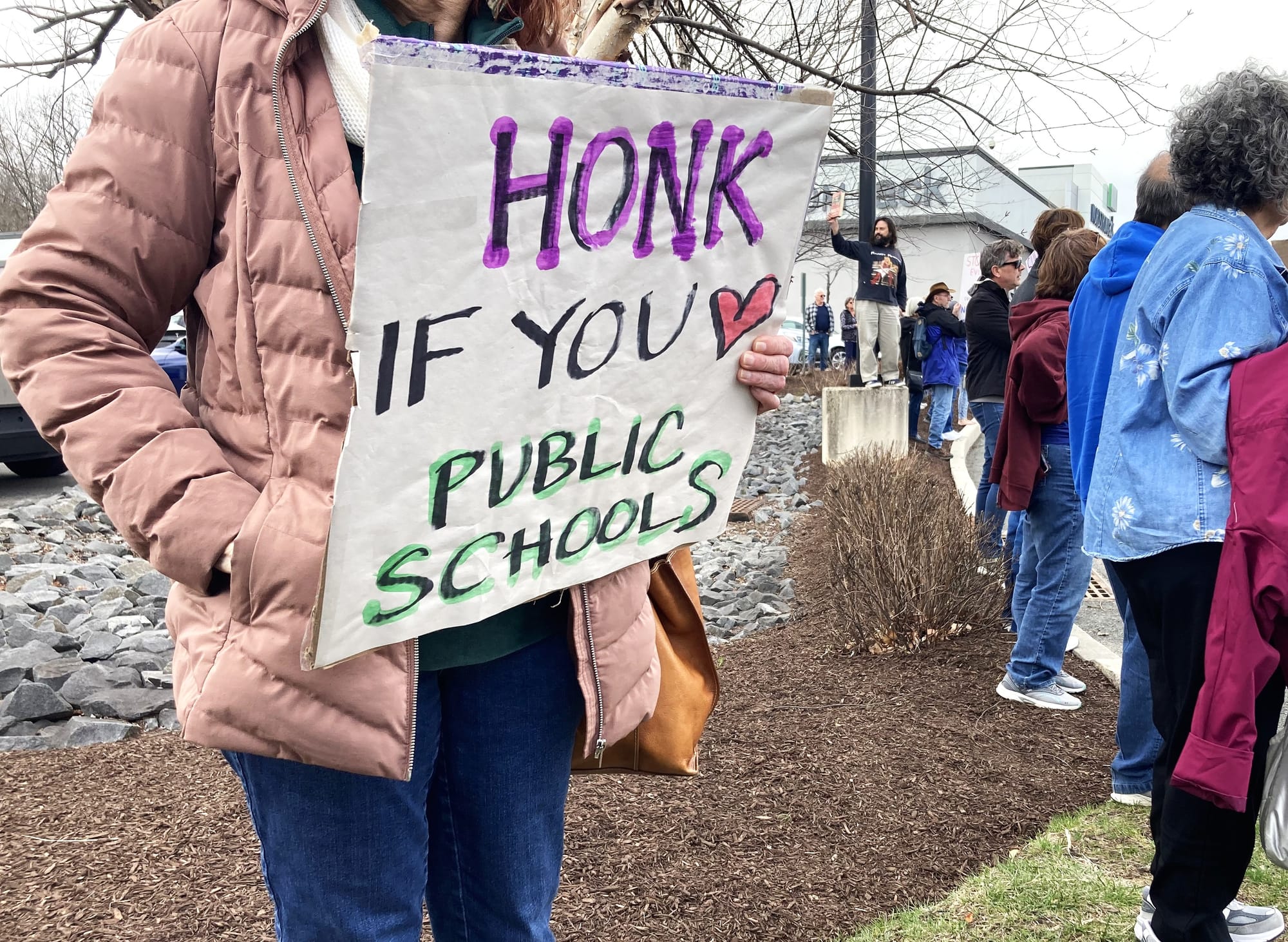 Close-up of a protester, face obstructed from view. They have one hand in a coat pocket and the other holds a HONK IF YOU HEART PUBLIC SCHOOLS sign.