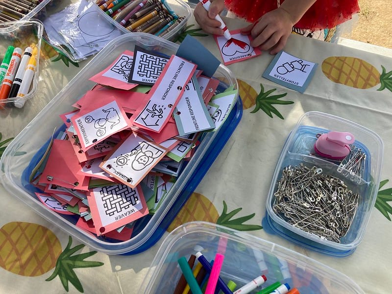 A child's hand coloring a little paper graphic with a heart on it. The social change role graphics are on paper cutouts alongside safety pins so that participants can pin their "badges" that show the roles they play on their clothing.