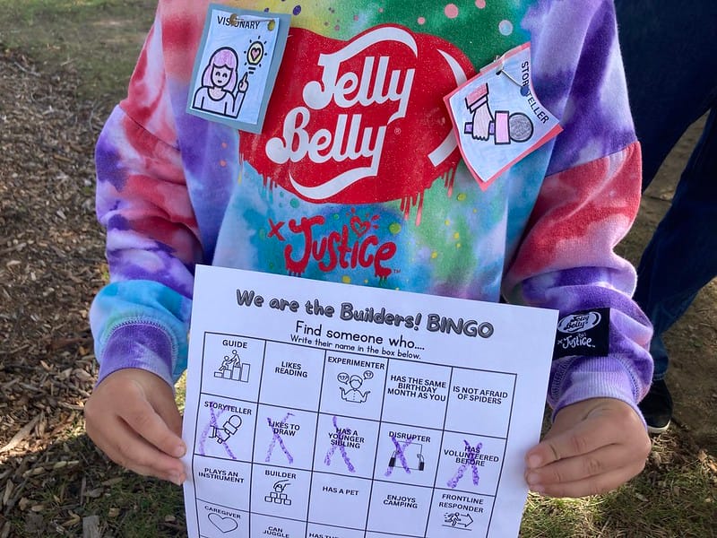 Child wearing a Jelly Belly tie-dyed sweatshirt and two We Are the Builders badges pinned onto it holds up their BINGO sheet with a whole row crossed out. Face not shown.