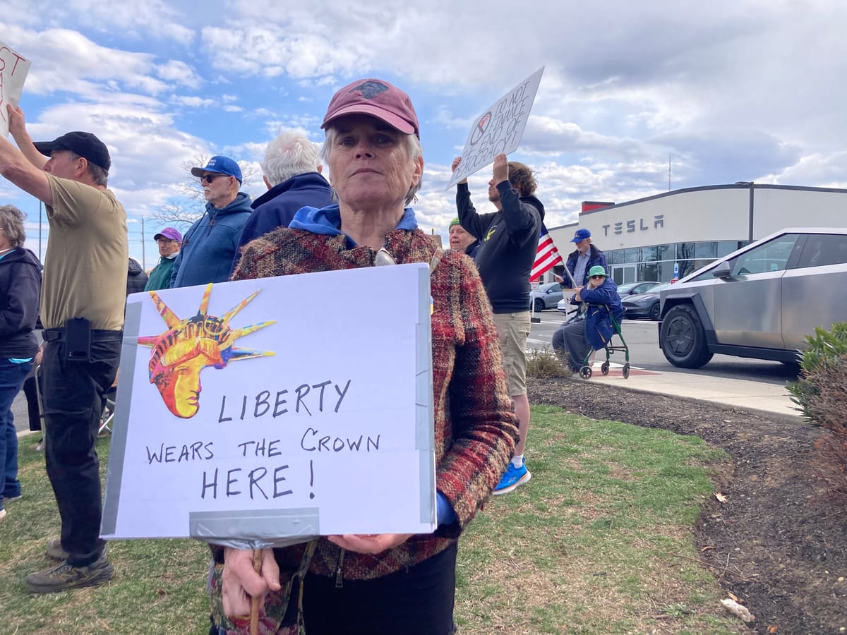 In front of a Tesla store, a middle-aged white person wears a baseball cap and serious expression. They hold a sign with a Statue of Liberty picture that reads LIBERTY WEARS THE CROWN HERE!