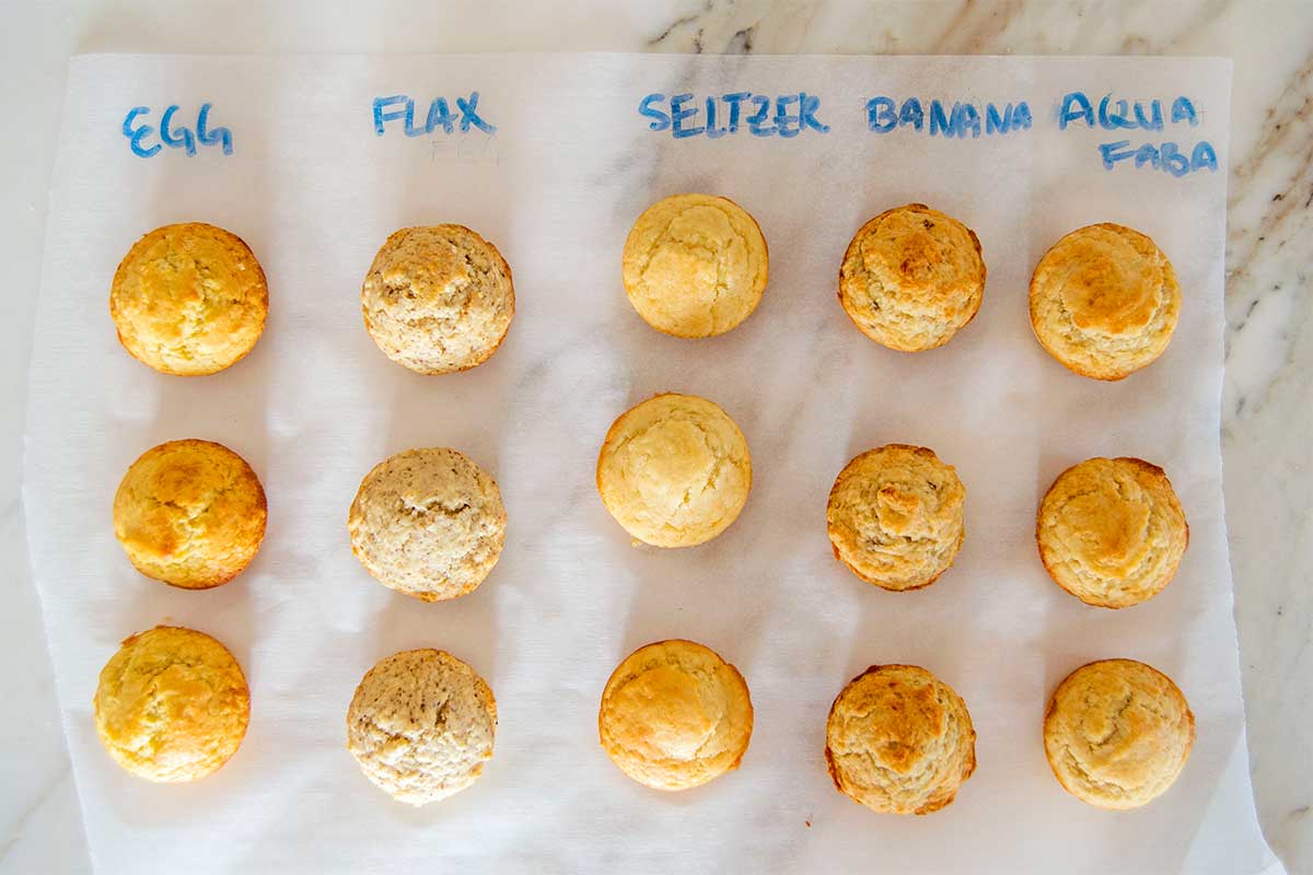 Photo of 5 different batches of muffins. In the left column are ones made with egg, then come Flax, Seltzer, Banana, and Aquafaba. Each has a slightly different golden color and rise.