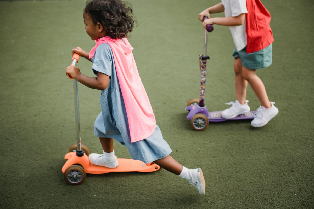 Two children, faces obscured, wearing makeshift superhero capes ride on small scooters on a dark green surface