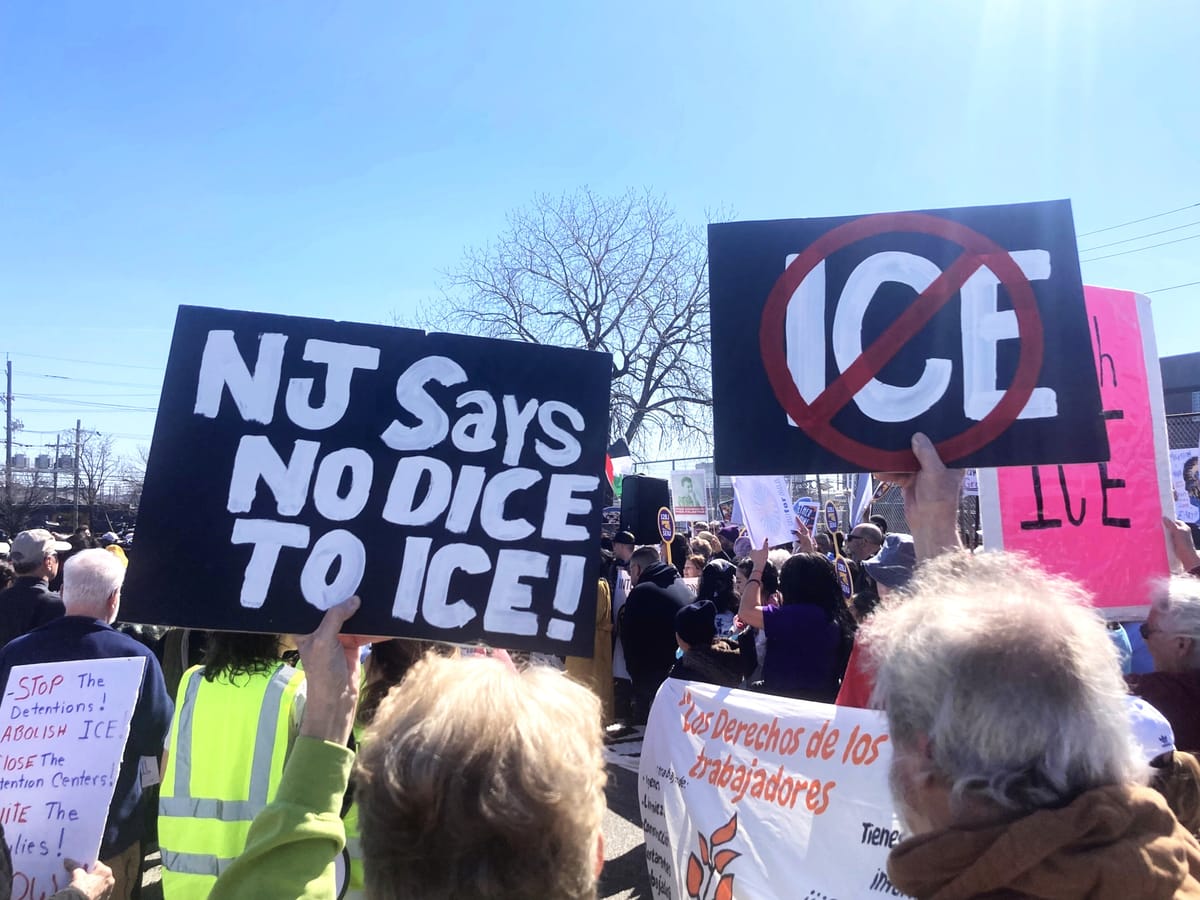 Under a blue sky, protesters hold up signs with anti-ICE logos, such as NJ SAYS NO DICE TO ICE!