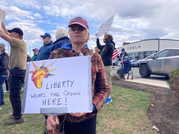 In front of a Tesla store, a middle-aged white person wears a baseball cap and serious expression. They hold a sign with a Statue of Liberty picture that reads LIBERTY WEARS THE CROWN HERE!