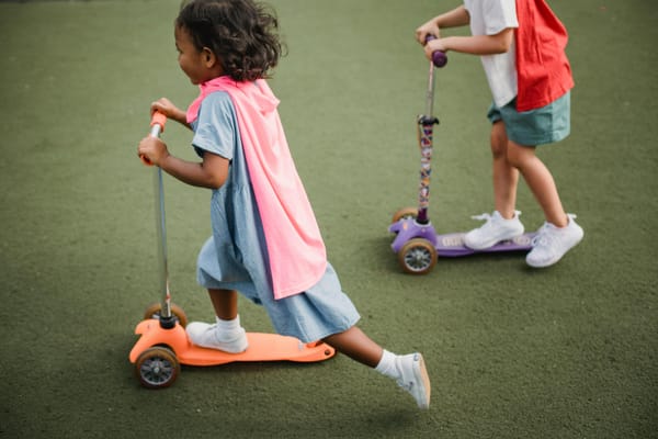 Two children, faces obscured, wearing makeshift superhero capes ride on small scooters on a dark green surface