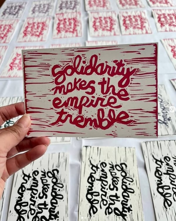 A hand holds a postcard-sized print that reads "solidarity makes the empire crumble" in linocut with red ink. Below, on a table, are many more of the prints in both black and red.