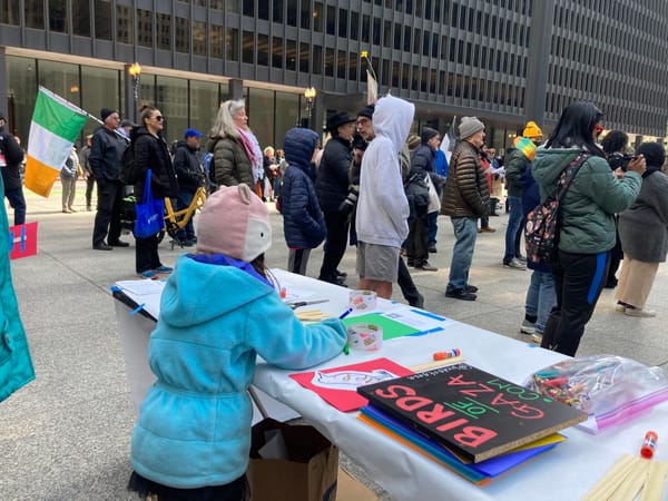 Near a large assembled crowd, a child facing away from the camera colors a bird illustration to dedicate to a child killed in Gaza.