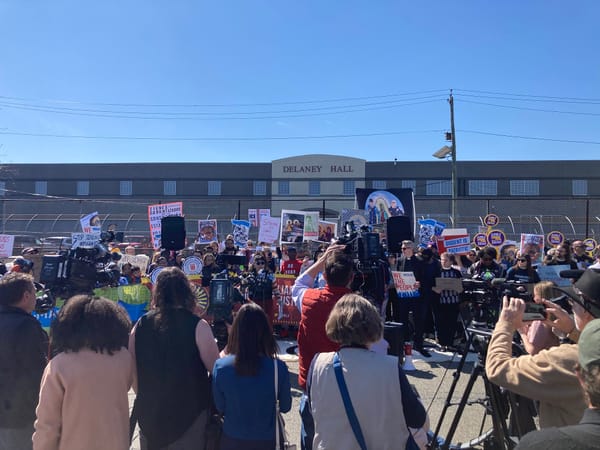 Under a cloudless blue sky, a crowd of ppl holding signs, news cameras, and microphones, outside a giant industrial building with DELANEY HALL above the entrance.