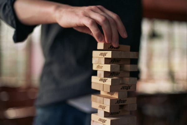Close-up of a person's slim brown hand gently placing a Jenga block atop a precariously-balanced tower