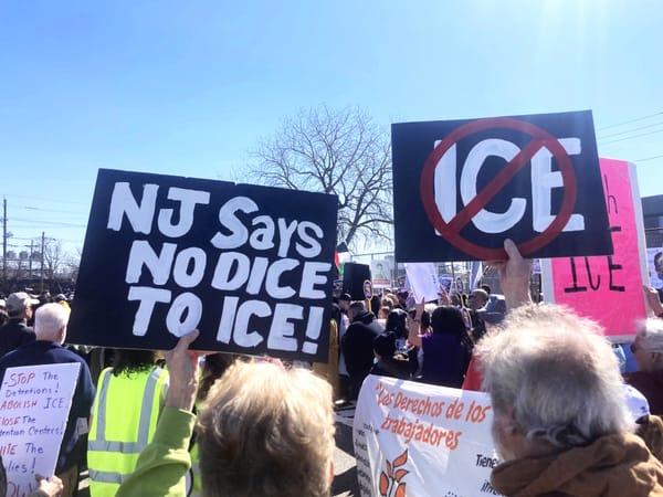 Under a blue sky, protesters hold up signs with anti-ICE logos, such as NJ SAYS NO DICE TO ICE!