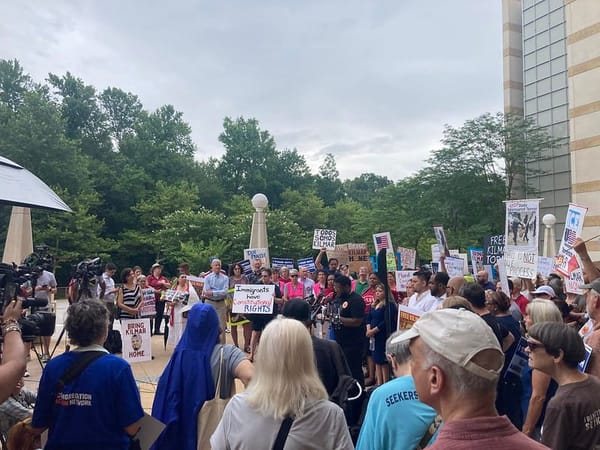 Large crowd gathered under cloudy skies. Handmade signs have slogans like IMMIGRANTS HAVE CONSTITUTIONAL RIGHTS and BRING KILMAR HOME. A rep from BAJI speaks at a mic.